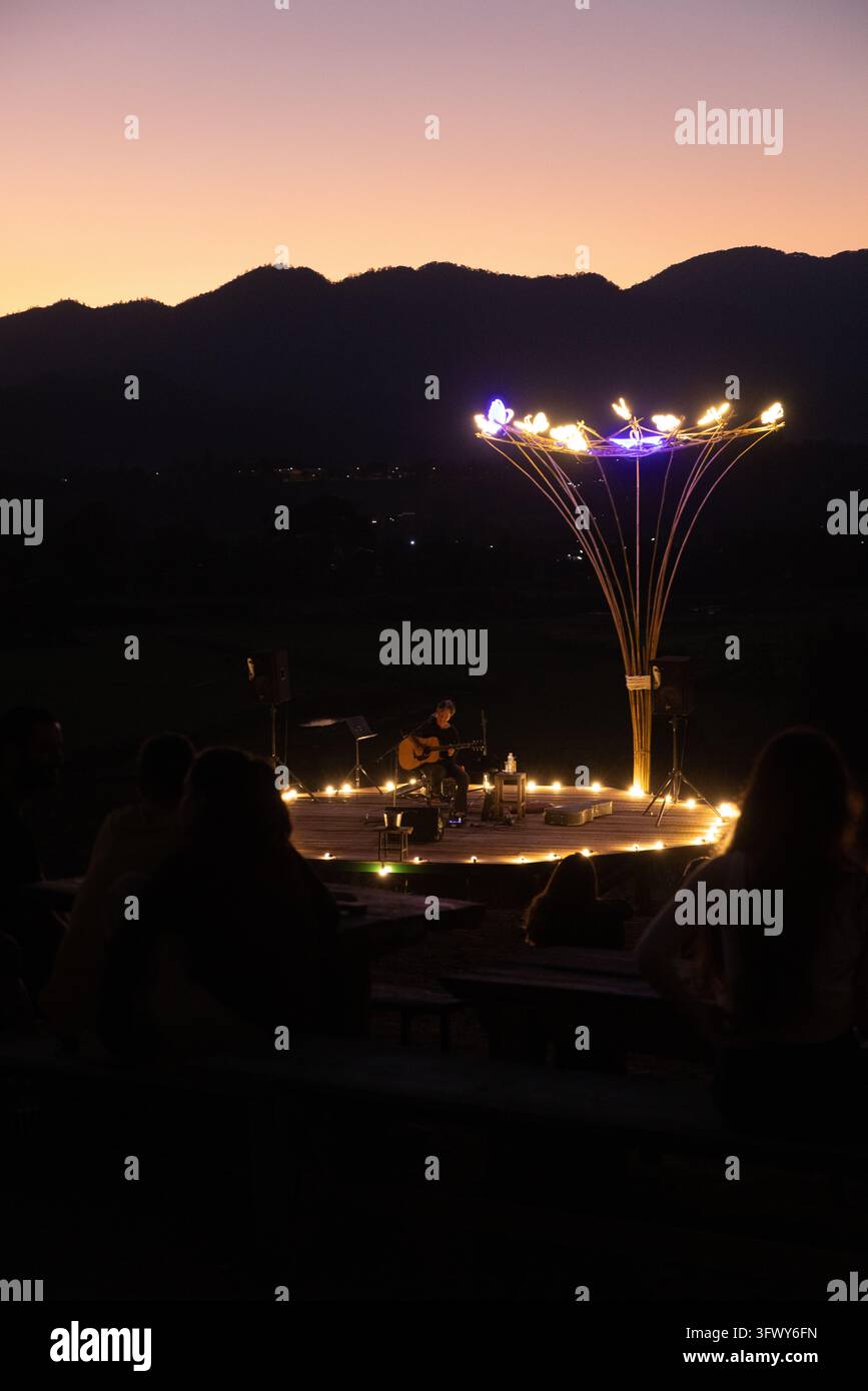 Pai, Thailandia - 20 gennaio 2026: Un musicista suona la chitarra su un palco circolare in legno illuminato da luminarie e un albero luminoso decorativo... Foto Stock