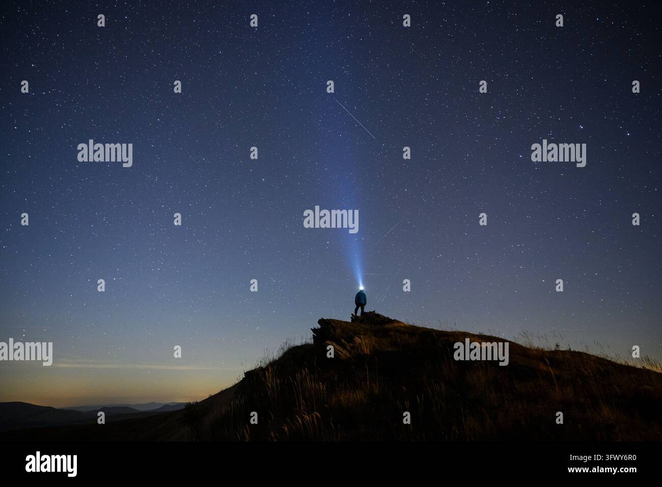 Un uomo solitario si trova su una collina rocciosa sotto un cielo notturno stellato con un fascio di luce brillante che si innalza verso l'alto. Una silhouette scura e un orizzonte lontano creano una scena astronomica drammatica Foto Stock