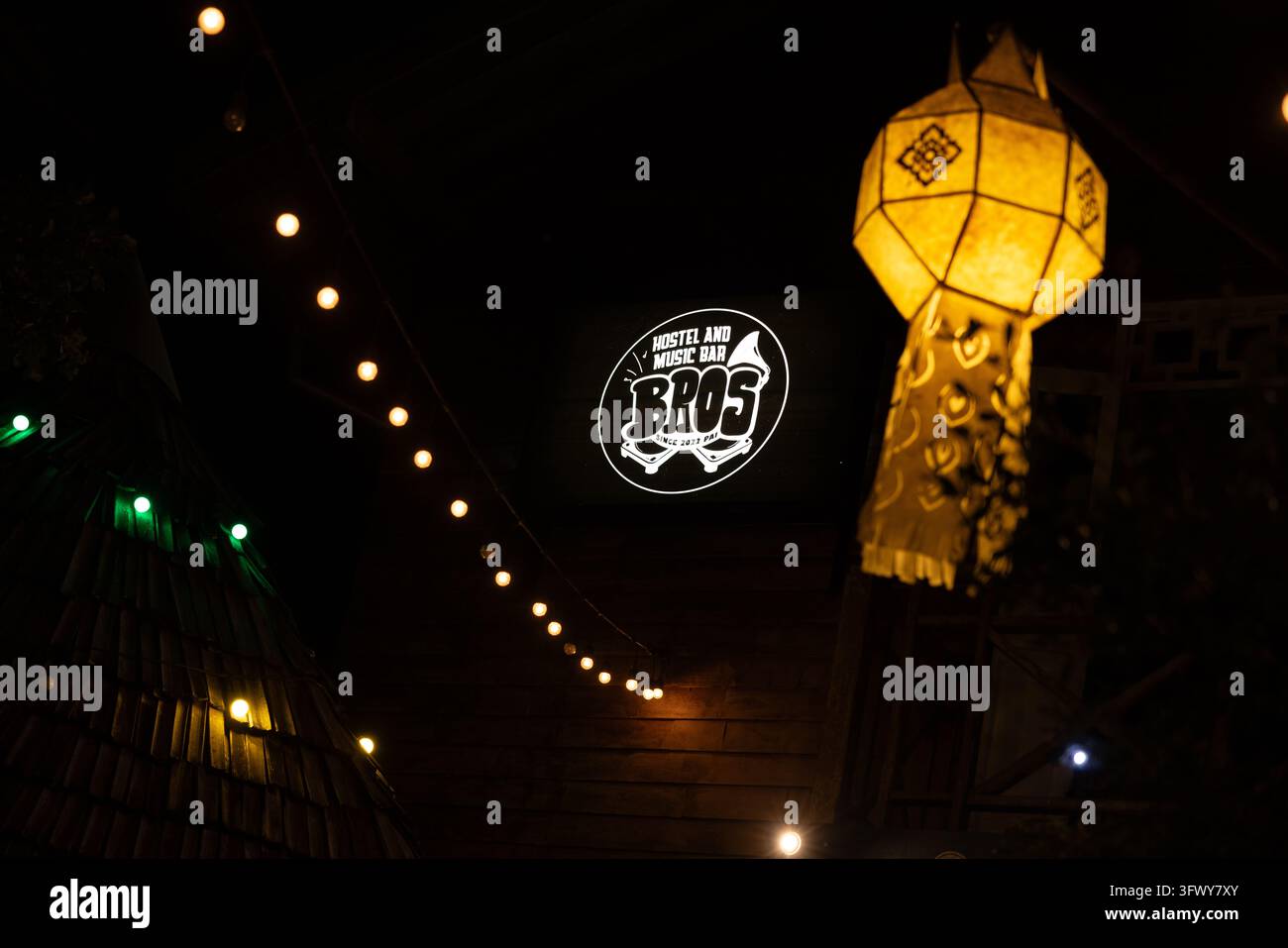 Pai, Thailandia - 22 gennaio 2026: Una lanterna luminosa pende accanto a una stringa di luci che illuminano il logo circolare di Bros Hostel e Music Bar... Foto Stock