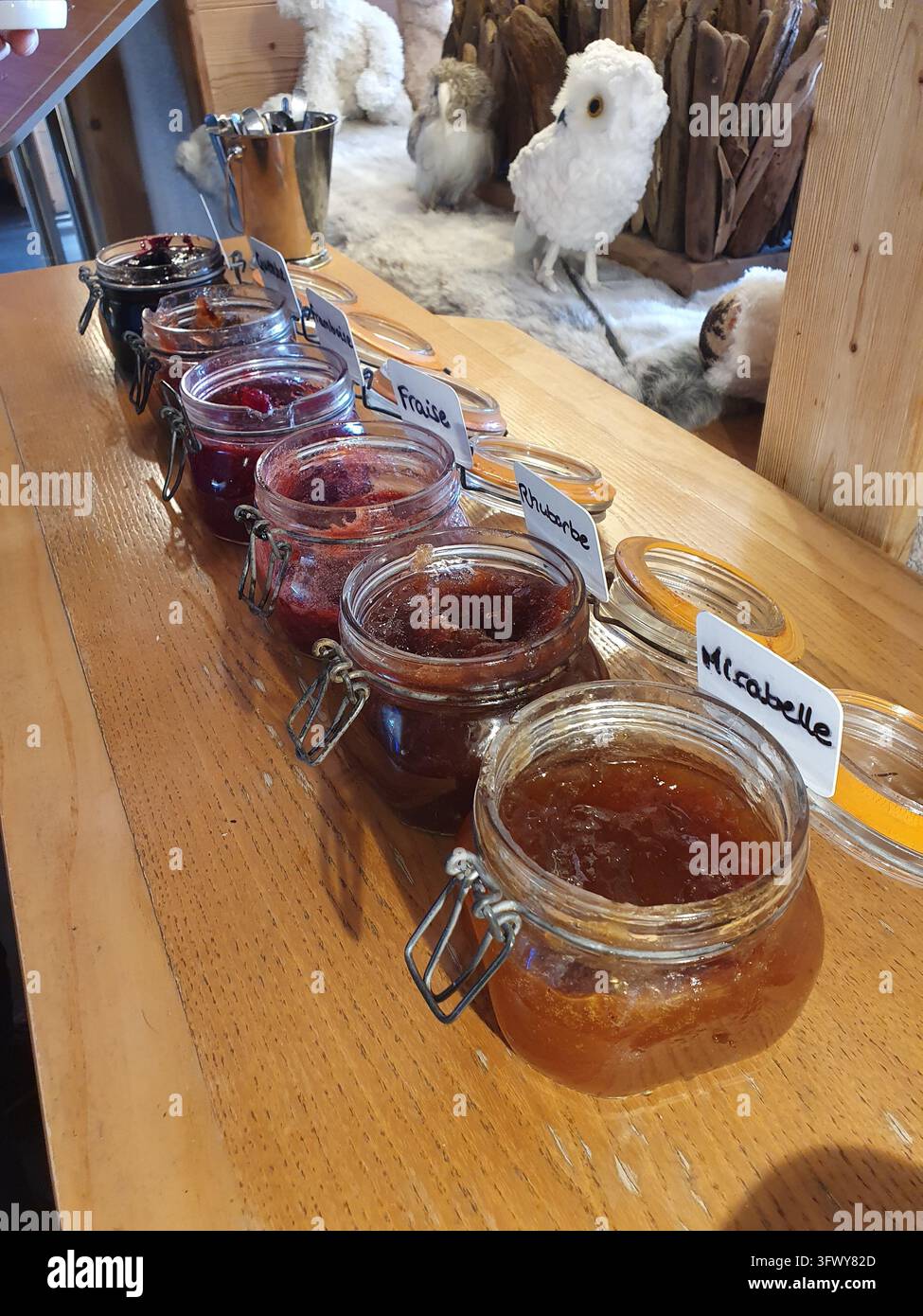 Diversi barattoli di marmellate e gelée di frutta assortite allineati su un bancone di legno rustico con etichette scritte a mano, creando una colazione artigianale e accogliente Foto Stock