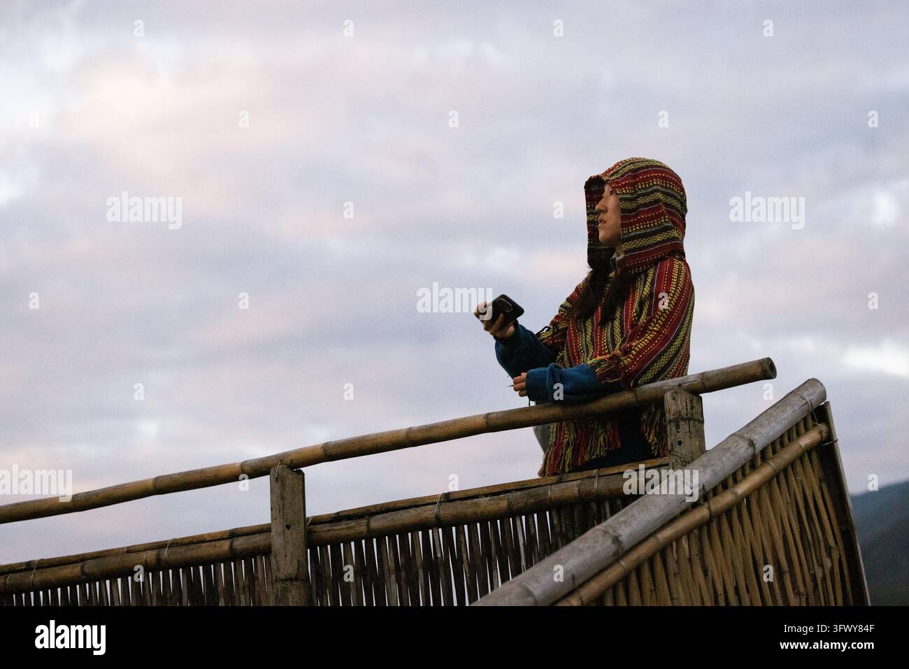 Pai, Thailandia - 23 gennaio 2026: Una donna che indossa uno scialle a strisce colorato con cappuccio si trova su un balcone di bambù rustico tenendo un telefono contro un... Foto Stock