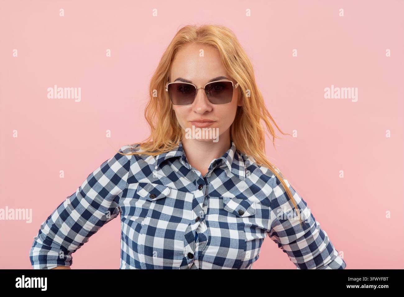 Donna bionda che posa con le mani sui fianchi, indossa una camicia a quadri e occhiali da sole, mostrando un'espressione sicura e seria su uno sfondo rosa Foto Stock