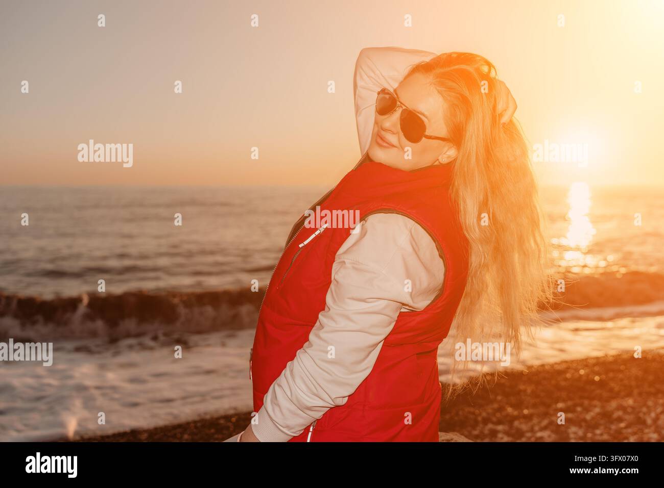 Donna spiaggia tramonto donna bionda in gilet rosso in posa su una spiaggia di ciottoli durante l'ora dorata Foto Stock