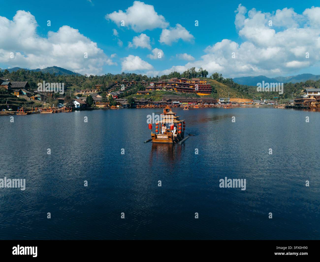 Vista aerea di una barca tradizionale in legno con lanterne rosse su un lago calmo a Ban Rak Thai, circondata da piantagioni di tè sulle colline e paesaggi montani Foto Stock