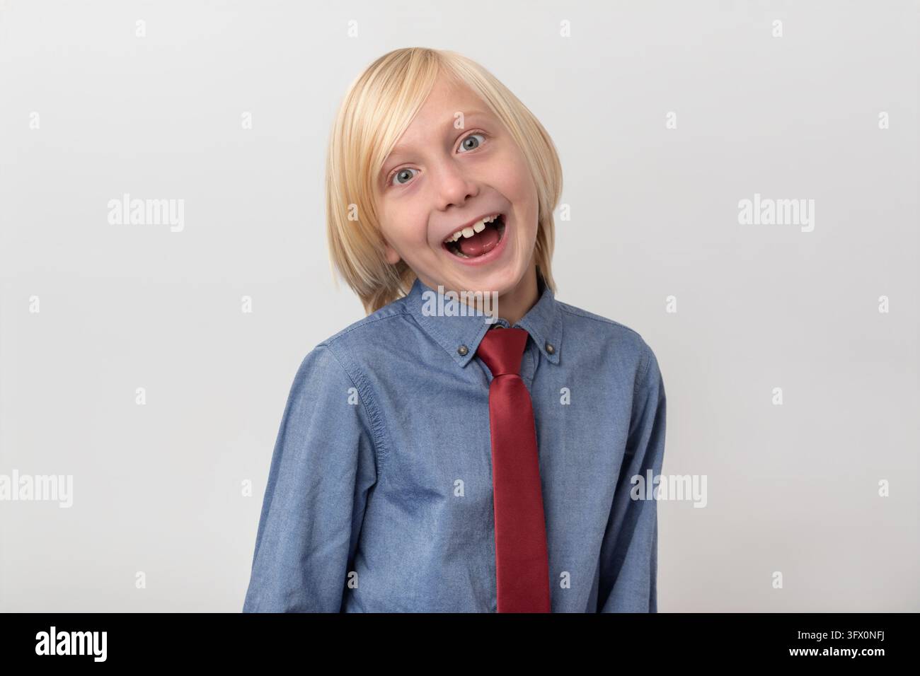 Ragazzo felice con capelli biondi e una cravatta rossa che guarda l'obiettivo con un sorriso a bocca aperta, esprimendo gioia e sorpresa Foto Stock