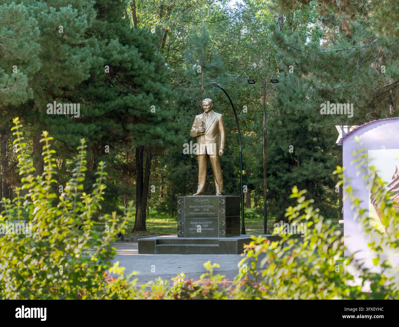 Statua dorata di Atatürk su un piedistallo di granito scuro tra i pini nel Parco Atatürk, Bishkek, Kirghizistan, incorniciata da arbusti a foglia larga. Foto Stock
