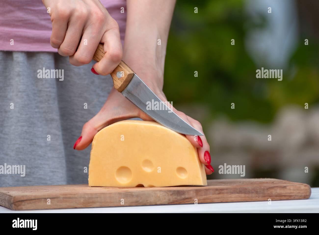 Mani di una donna con unghie rosse che tagliano un grande blocco di formaggio svizzero giallo con un coltellino su un tagliere di legno, primo piano della preparazione del cibo Foto Stock