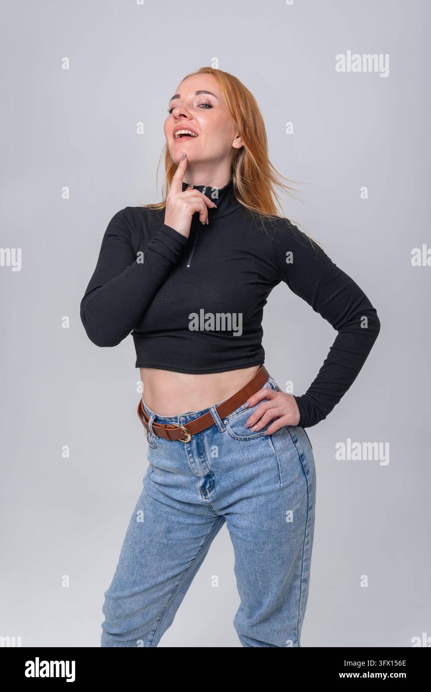 Giovane donna dai capelli rossi in abbigliamento casual elegante posa con la mano sul mento, guarda verso l'alto con un sorriso sottile, trasmettendo sicurezza riflessiva, creatività e Foto Stock