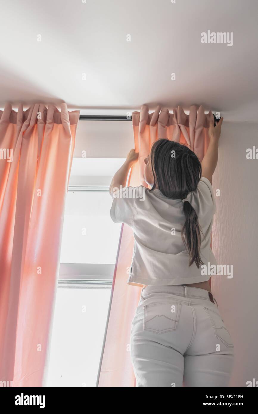 Vista posteriore di una donna con capelli scuri raccolti in una coda di cavallo, che indossa una maglietta bianca e jeans chiari, che si allunga verso l'alto per sistemare tende rosa su un'asta nera Foto Stock