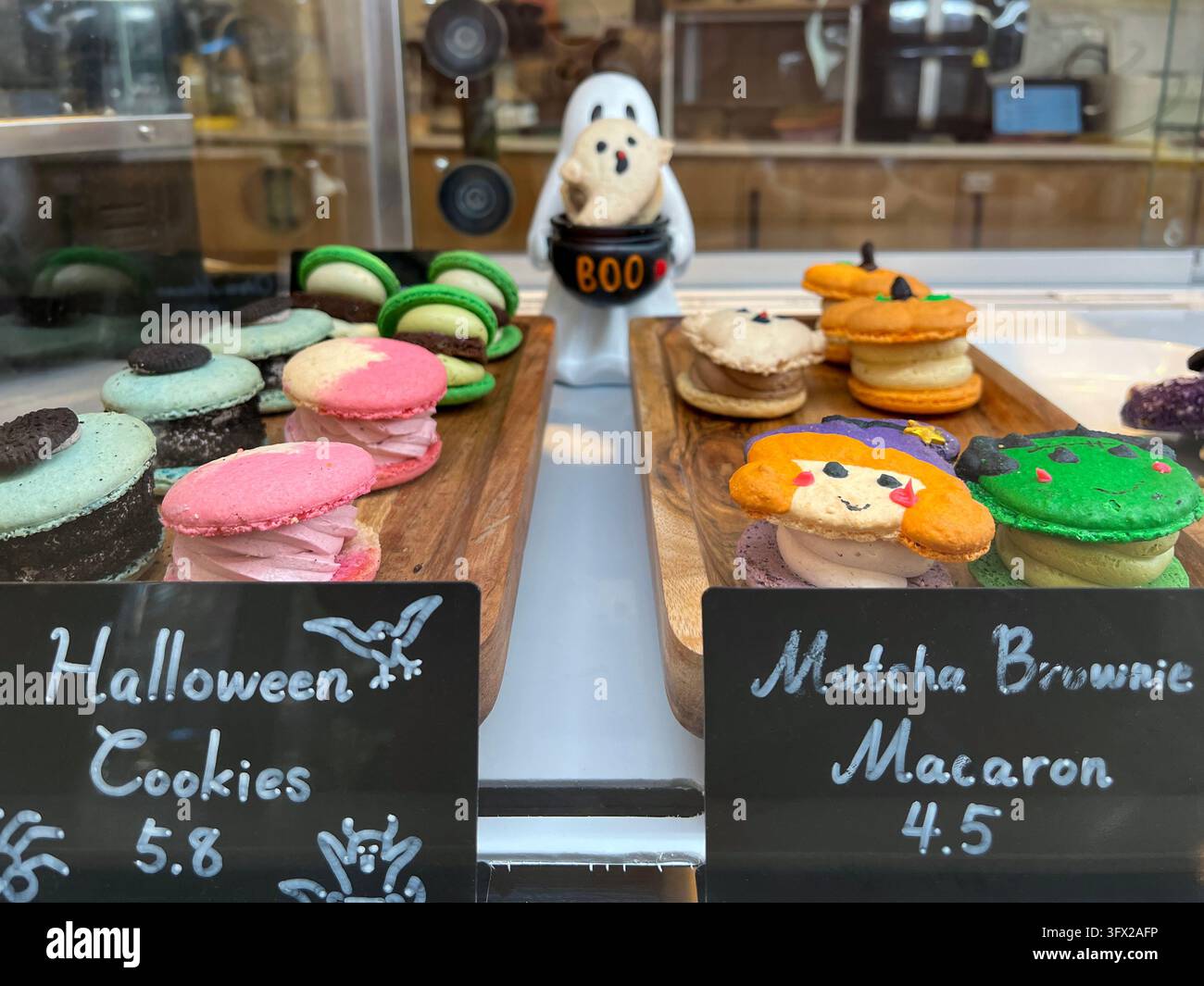 Insegne in lavagna con scritto Biscotti di Halloween e Macaron al Brownie Matcha all'interno di una vetrina di pasticceria piena di pasticcini festivi e una statuetta di fantasma Foto Stock