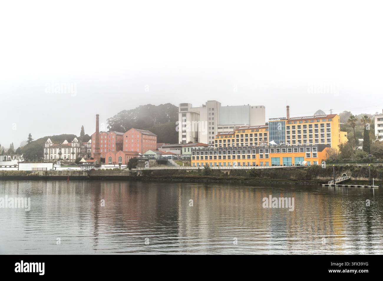 Edifici industriali rosa con un camino in mattoni, un palazzo bianco e un hotel giallo si trovano sul Douro tra Porto e Vila Nova de Gaia, Portogallo. La nebbia aleggia sulla scena. La scritta "Pestana Douro" è presente sull'hotel. Un alto edificio di colore chiaro si erge dietro. I colori si riflettono nell'acqua calma. Foto Stock