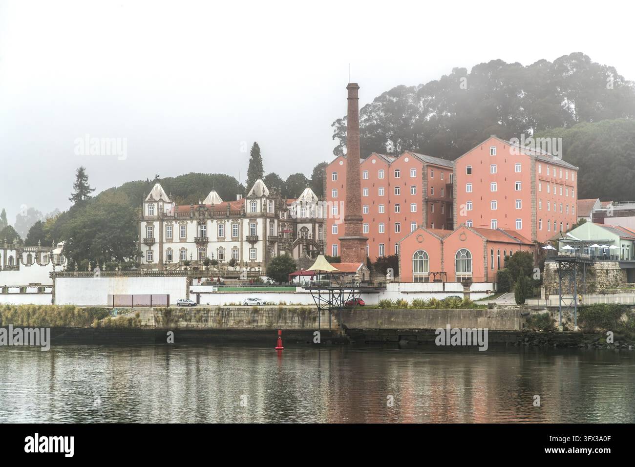 Un palazzo bianco con torri angolari e tetti appuntiti si trova accanto ad edifici industriali rosa con un camino in mattoni sul Douro tra Porto e Vila Nova de Gaia, Portogallo. Il palazzo potrebbe essere il Palácio do Freixo (Palazzo di Freixo). Una boa rossa galleggia nel fiume. La nebbia nasconde le colline sullo sfondo. Foto Stock