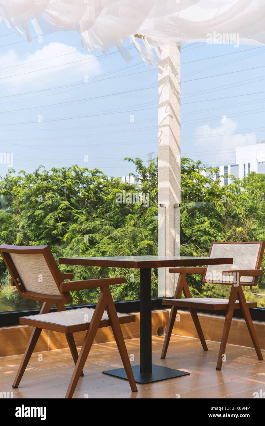 Area di seduta minimalista moderna di un caffè con due sedie di legno e un tavolo quadrato su un balcone illuminato dal sole che si affaccia su alberi verdi rigogliosi e lo skyline urbano sotto un baldacchino bianco trasparente, caratterizzato da linee pulite, mobili in teak dai toni caldi, schienali in vimini intrecciati, base in metallo nero, pavimento piastrellato e linee elettriche sopra la testa contro un cielo blu brillante con nuvole sparse Foto Stock