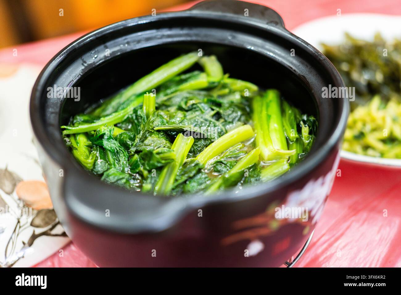 Spinaci al vapore in una tradizionale casseruola di ceramica nera, serviti caldi con brodo chiaro, tipici della cucina casalinga cinese; sullo sfondo si vedono contorni complementari tra cui verdure sottaceto e aglio tritato saltato su una tovaglia rossa. Foto Stock