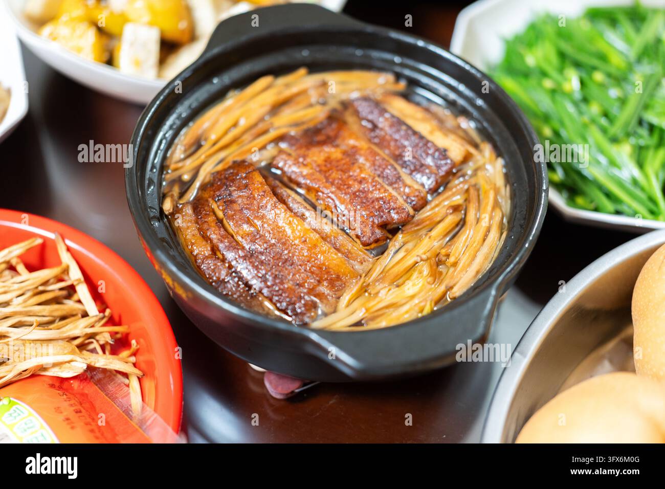 Anatra stufata con funghi enoki in una pentola di ceramica nera, servita calda su un tavolo da pranzo in legno insieme a contorni tra cui tofu al vapore, scalogni freschi e verdure sottaceto, caratteristica della cucina tradizionale cinese del sud, probabilmente della provincia di Guangdong o Fujian Foto Stock