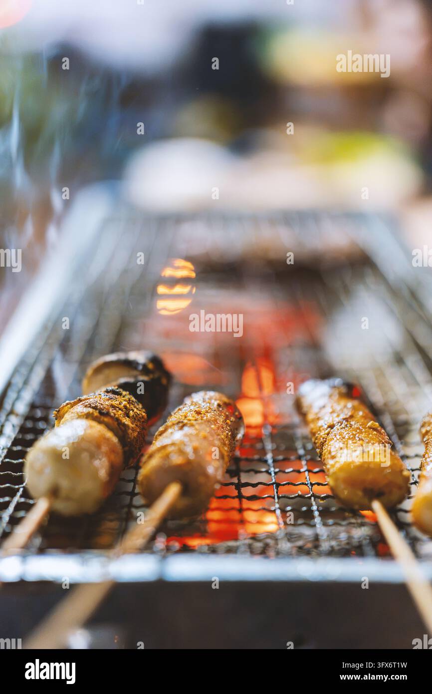 Spiedini di yakitori grigliati che sfrigolano su carboni ardenti su una griglia metallica tradizionale, con pezzi di pollo conditi con una grigliatura dorata e fili di fumo che si alzano, catturati con una profondità di campo poco profonda e illuminazione ambientale calda tipica di un izakaya di Tokyo o di un chiosco da strada Foto Stock