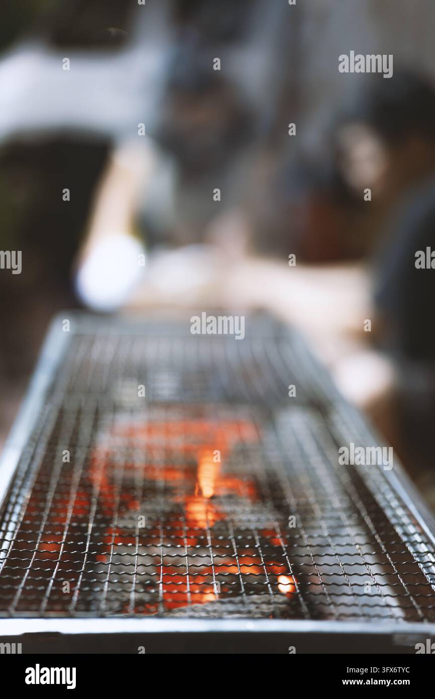 Primo piano di una tradizionale griglia giapponese yakiniku con braci di carbone incandescenti sotto una griglia metallica, commensali sfocati sullo sfondo che suggeriscono un'esperienza culinaria indoor intima in un izakaya di Tokyo o in un ristorante specializzato in carni Foto Stock