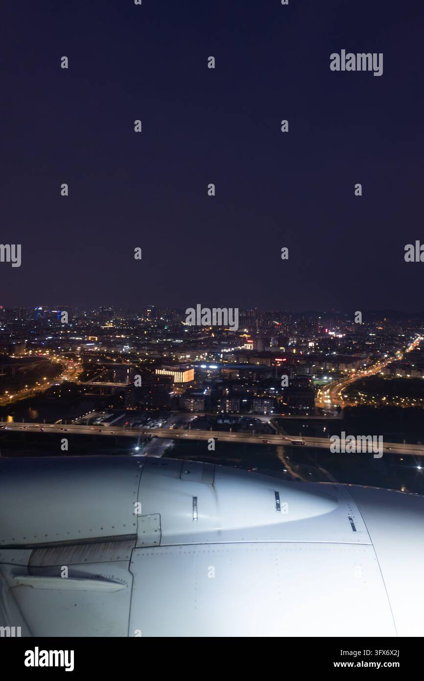 Vista aerea da un finestrino dell'aereo di notte che mostra l'ala illuminata in primo piano e un'estesa veduta urbana sottostante con strade, edifici e reti autostradali luminose, probabilmente scattata durante l'avvicinamento o il decollo su una grande metropoli est-asiatica come Shanghai o Guangzhou, sotto un cielo scuro e sereno con minima copertura nuvolosa. Foto Stock