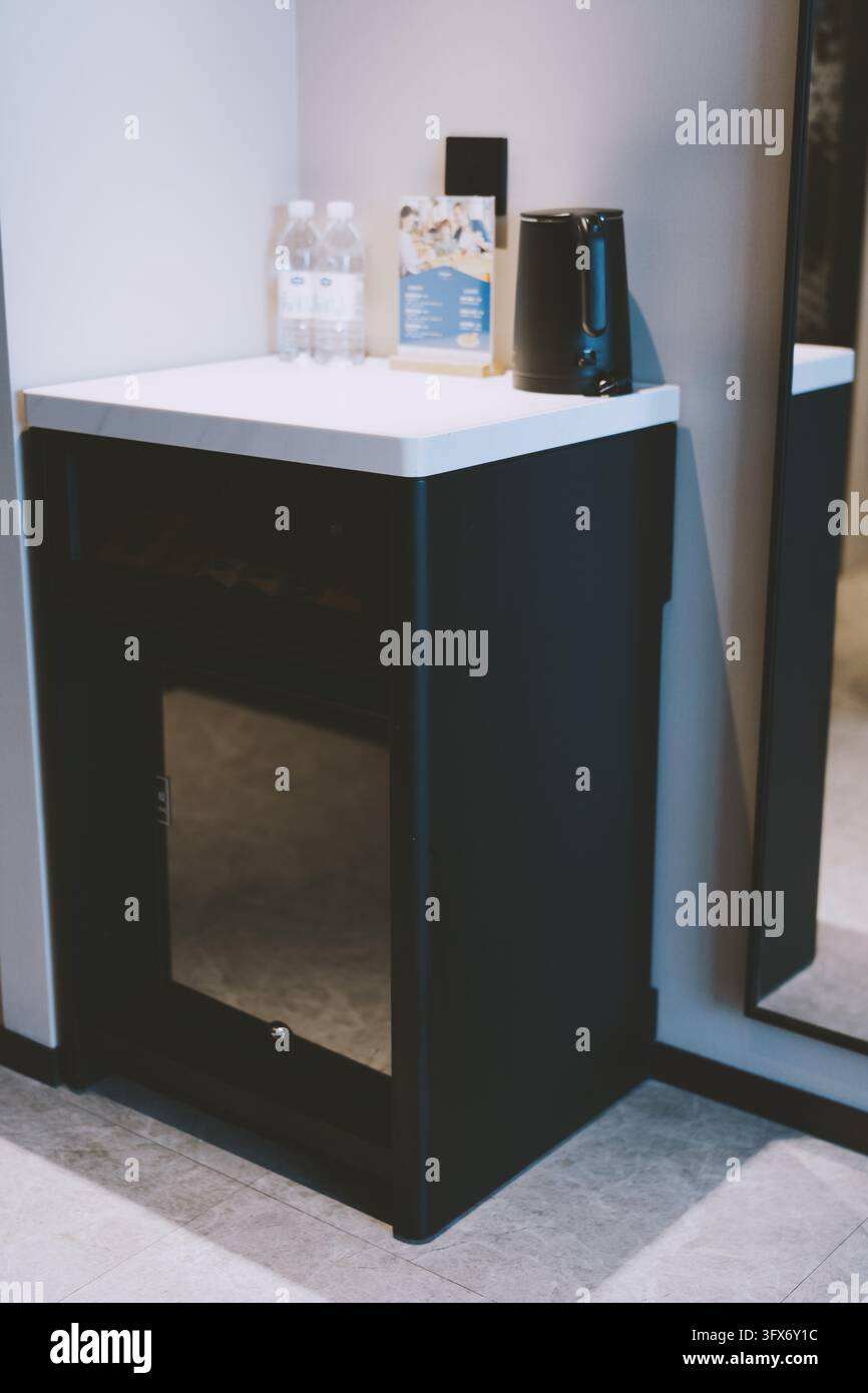 Moderno mini frigorifero nero con porta in vetro e piano di lavoro in marmo bianco nell'angolo di una camera d'albergo, sormontato da due bottiglie d'acqua di plastica, un bollitore elettrico nero e una brochure piegata, che riflette il design contemporaneo dell'ospitalità e la comodità degli ospiti Foto Stock
