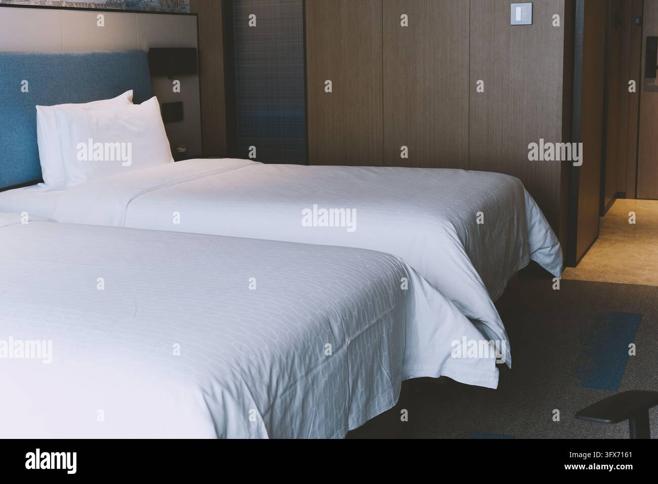 Camera d'albergo moderna con due letti singoli ordinatamente rifatti con biancheria a righe bianche, una testiera in tessuto blu strutturato, pannelli murali in legno scuro e illuminazione ambientale; design di interni contemporaneo per l'ospitalità in un contesto di alloggio urbano Foto Stock