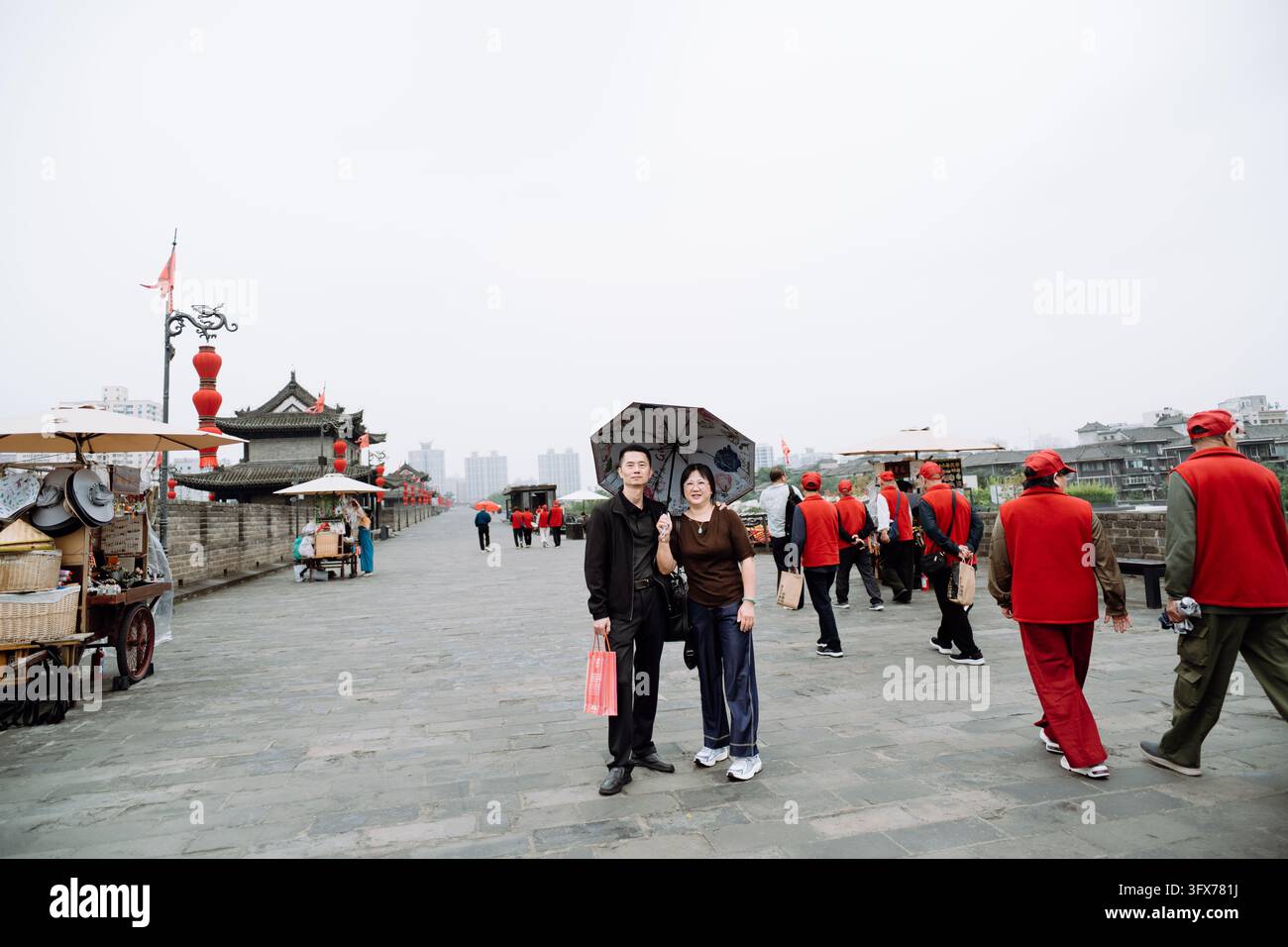 Una coppia sorridente di mezza età posa per una foto sul muro storico della città di Xi'an nella provincia dello Shaanxi, in Cina, tenendo un ombrello a motivi e borse della spesa, con architettura tradizionale cinese, lanterne rosse, bancarelle di souvenir e gruppi di turisti e volontari in giacche rosse che camminano lungo l'ampio marciapiede di pietra sotto un cielo nuvoloso Foto Stock