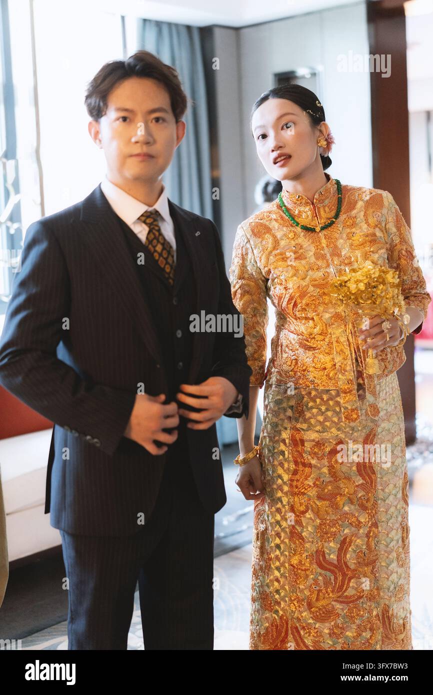 Una sposa asiatica elegante in un lussuoso qipao di seta dorata con intricati ricami di fenici e fiori, che indossa una collana di giada e tiene un delicato bouquet giallo, si trova accanto al suo sposo in un elegante completo a tre pezzi con gessato e cravatta a motivi, entrambi posano con sicurezza in un moderno atrio d'hotel con grandi finestre e luce naturale soffusa, catturando una fusione contemporanea del patrimonio cinese e dell'abbigliamento formale occidentale per un servizio fotografico pre-matrimonio a Shanghai Foto Stock