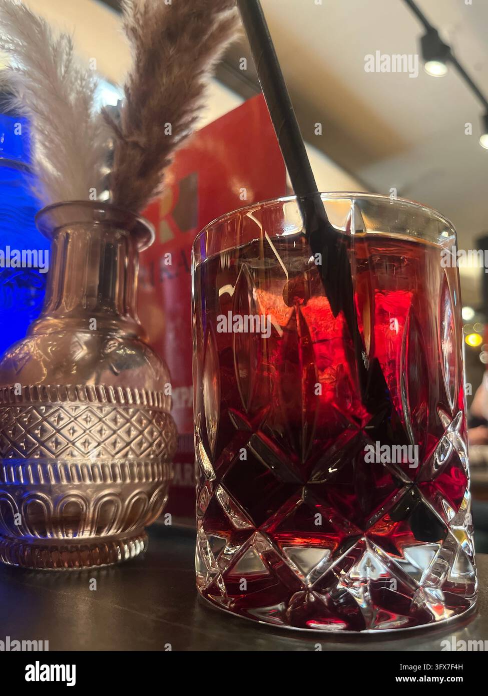 Un primo piano di un cocktail rosso scuro servito in un bicchiere di cristallo con una cannuccia nera.Illuminazione d'atmosfera cupa del bar.Vaso decorativo con erba. Foto Stock