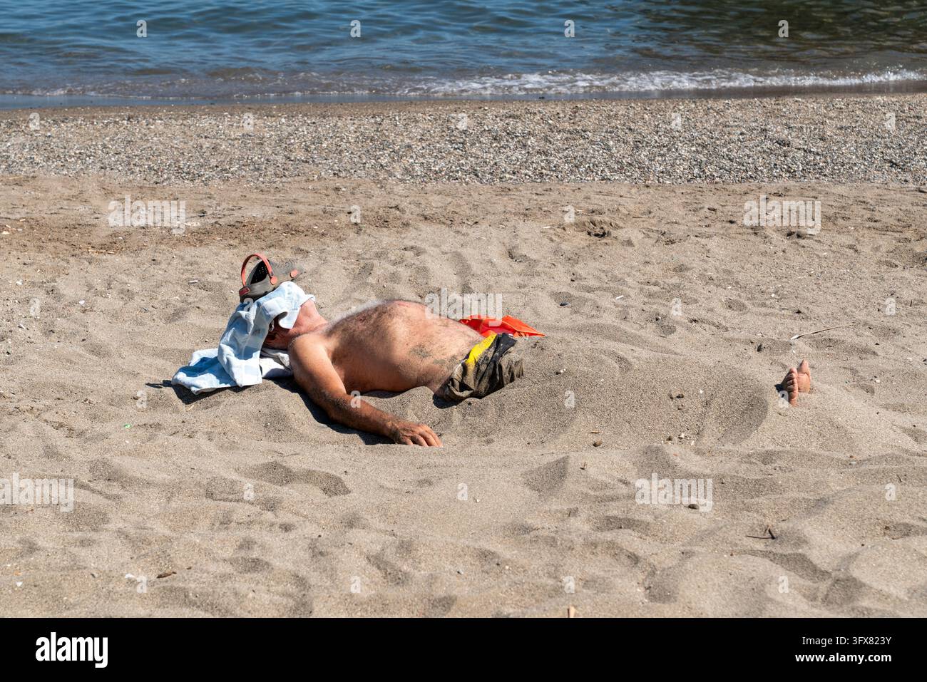 Un uomo anziano sepolto sotto la sabbia calda che si rilassa e fa il bagno al sole su una spiaggia di sabbia sul litorale marino. Volto coperto con un asciugamano e una ciabatta per l'ombra e pro Foto Stock
