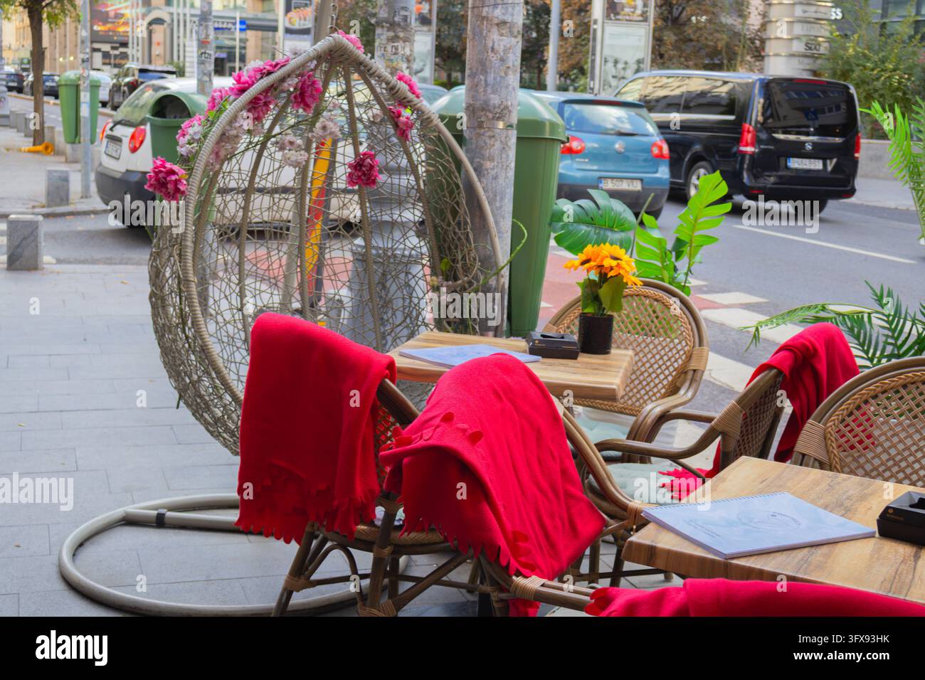 Sedie da caffè sul marciapiede con coperte rosso vivo, tavoli di legno e una sedia sospesa a forma di uovo decorata con fiori artificiali - Bucarest, Romania - ottobre Foto Stock