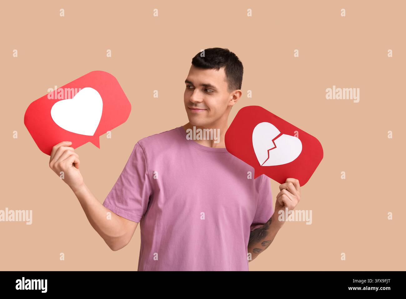 Incontri e scelte relazionali sui social media, giovane uomo che tiene icone di cuore e cuore spezzato che rappresentano l'amore e le decisioni di rottura su sfondo beige Foto Stock
