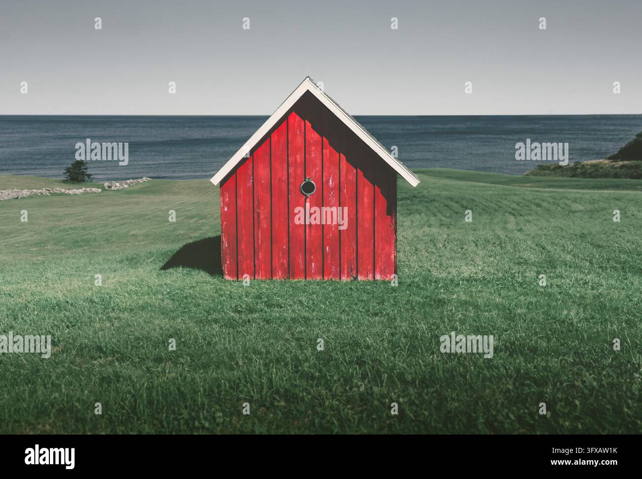 Un capannone di legno rosso si erge solo su un prato erboso accanto a un grande lago. La struttura semplice, la riva aperta e il cielo nuvoloso creano un paesaggio rurale tranquillo Foto Stock
