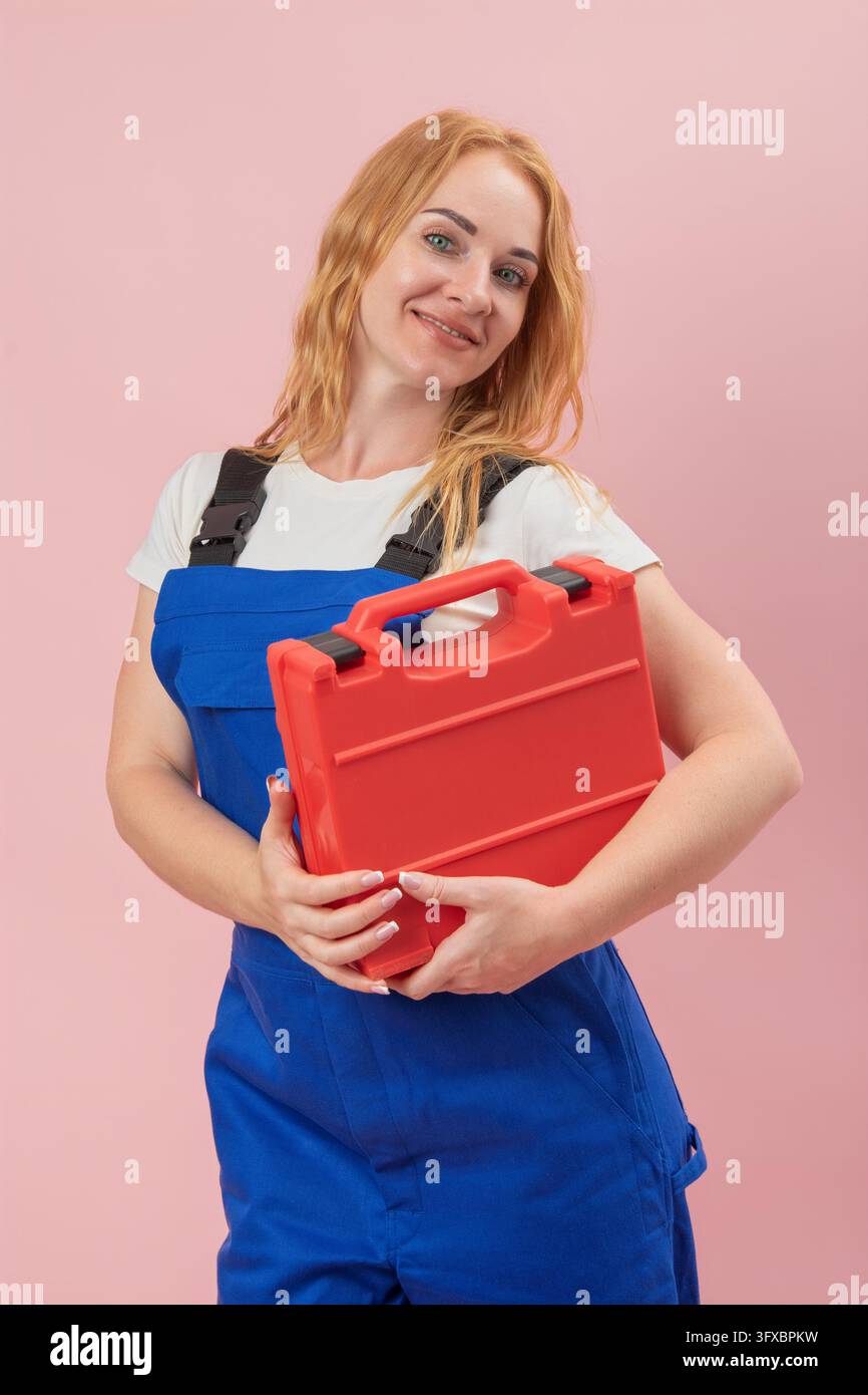Lavoratrice sicura di sé con tute blu professionali, sorride alla fotocamera e tiene una cassetta degli attrezzi rossa su uno sfondo rosa chiaro, rappresenta Foto Stock