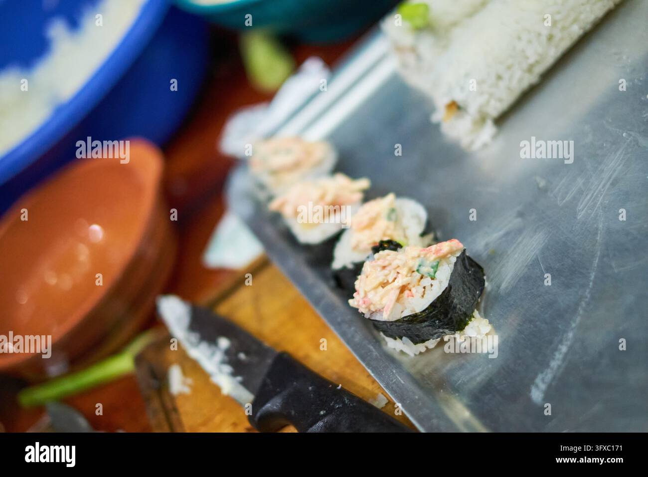 Mani che arrotolano e tagliano rotoli di sushi fatti in casa con nori e riso su una tavola di legno. Foto Stock