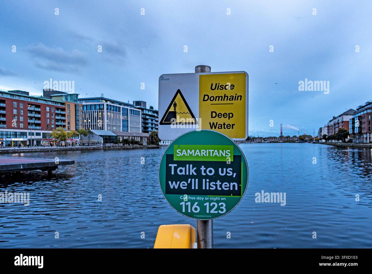 Segni di avvertimento per acque profonde e linea di assistenza Samaritans che sovrastano il Grand Canal Dock a Dublino, con edifici moderni sul lungofiume. Foto Stock