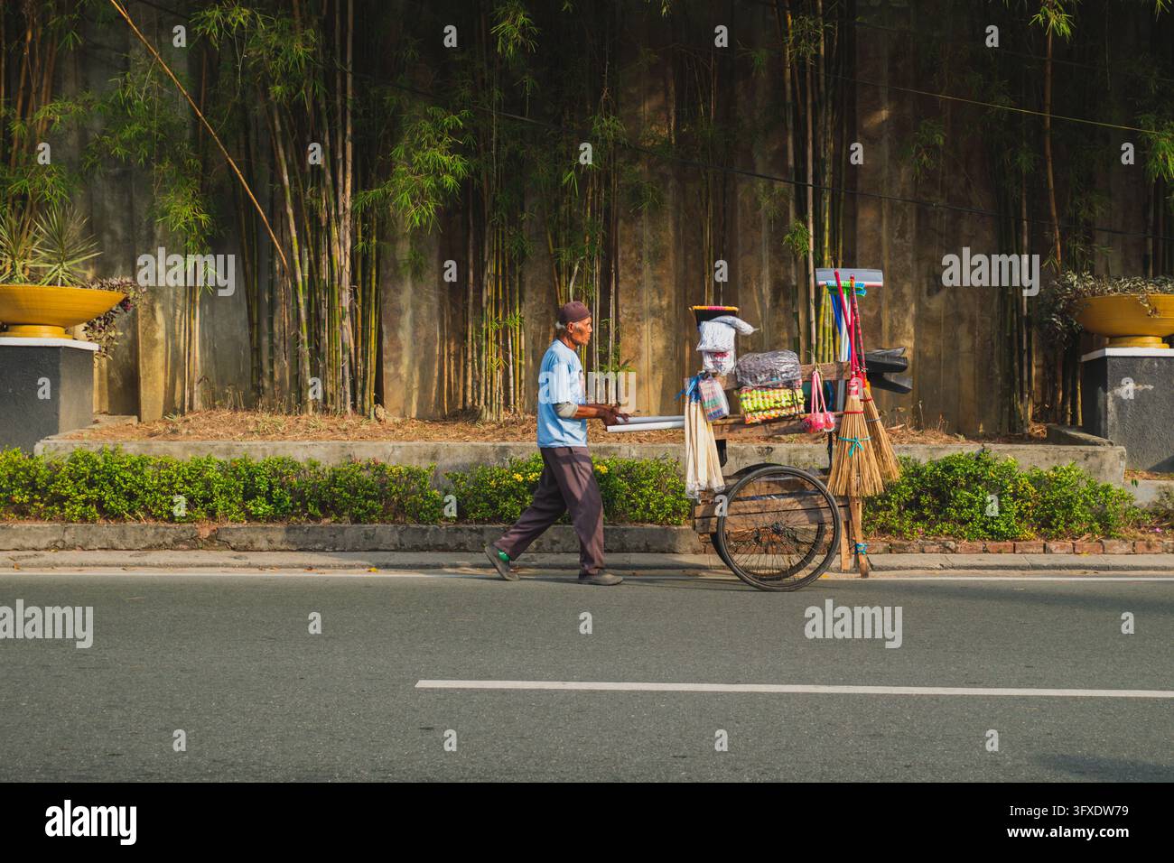 Balikpapan, Indonesia - 10 settembre 2026. Un venditore ambulante con una maglietta blu e pantaloni scuri spinge un carrello di legno carico di oggetti, scope e melma Foto Stock