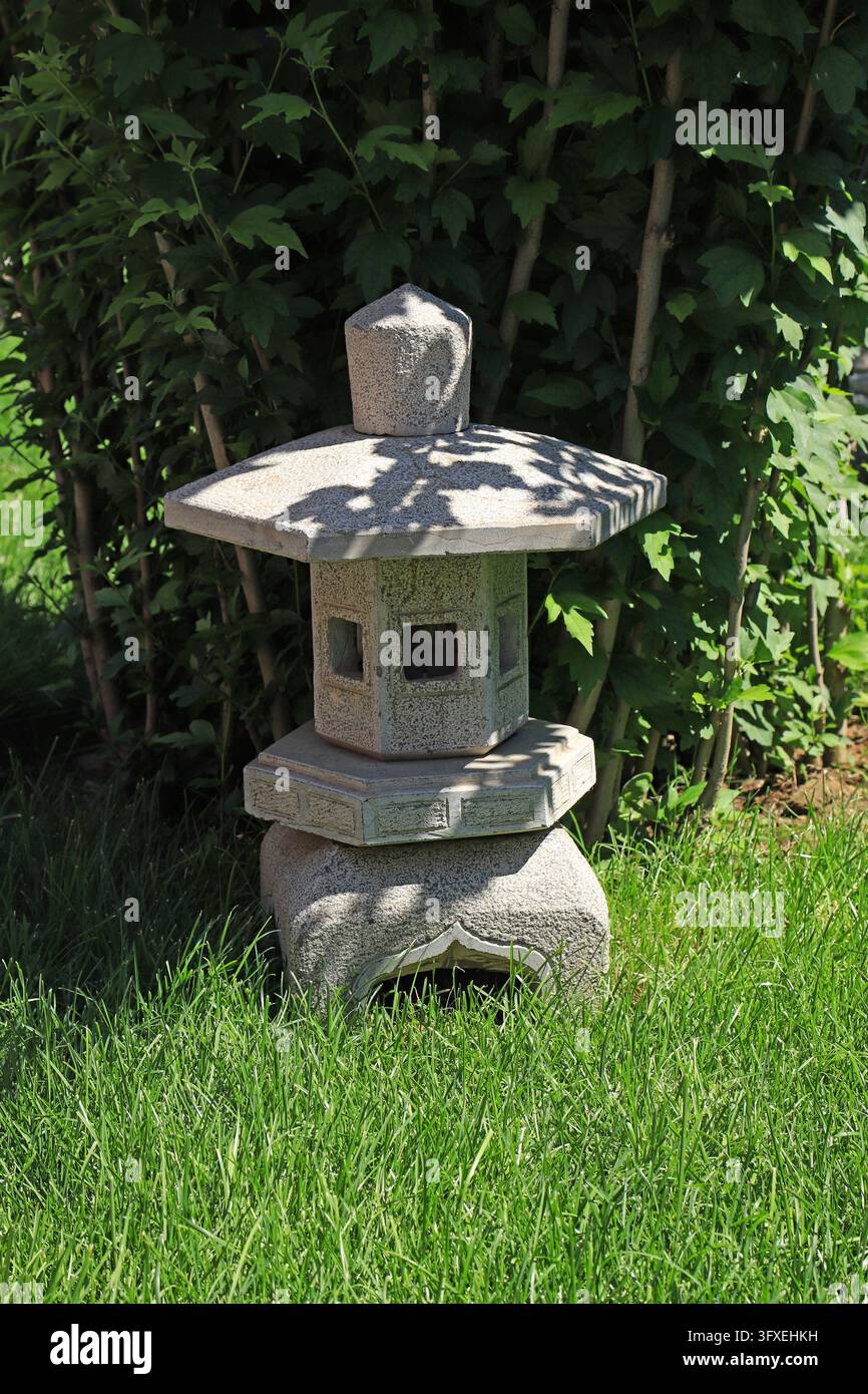 Antica lanterna in pietra blu in stile giapponese e cinese, posizionata su erba verde circondata da cespugli, decorazione paesaggistica zen per cortile, tè Foto Stock