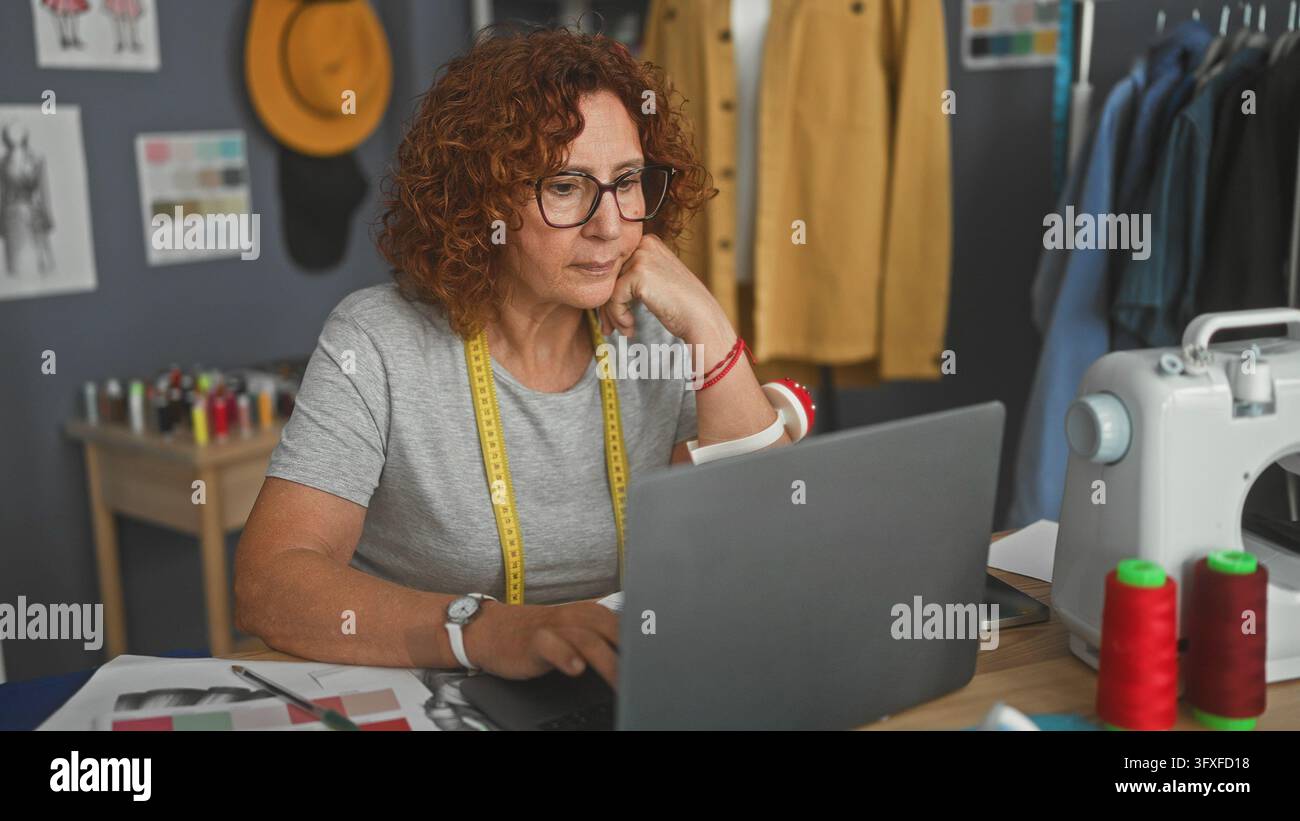 Sarta donna con metro a nastro davanti al laptop in studio, mano sul mento mentre lavora accanto alla macchina da cucire; concentrazione silenziosa. Foto Stock