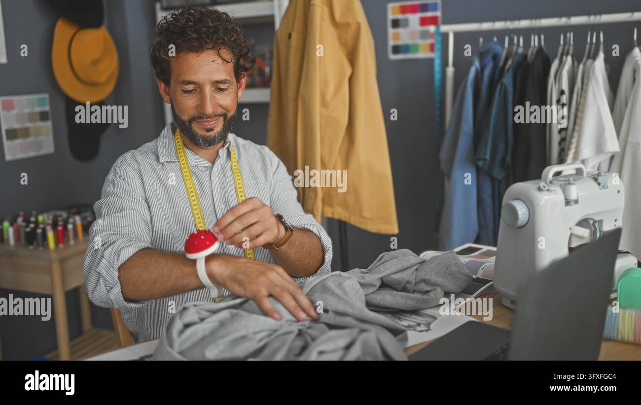 Uomo che cuce a mano un indumento grigio su un tavolo da studio con macchina da cucire, metro e laptop visibili; attenzione alla lavorazione artigianale. Foto Stock