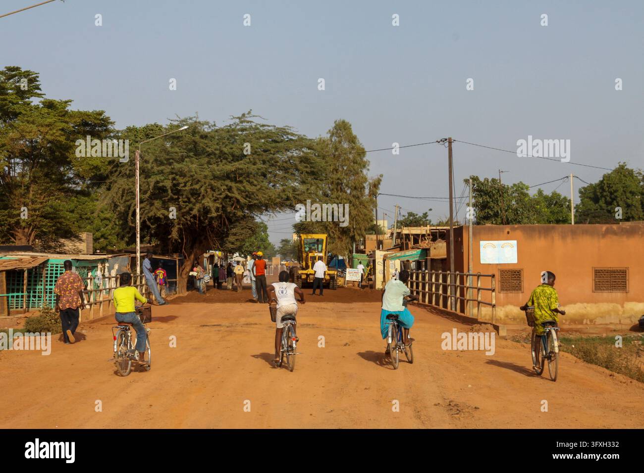 Persone in bicicletta su una strada sterrata fiancheggiata da alberi alti e edifici bassi, Regione Centro, Ouagadougou, Burkina Faso Foto Stock