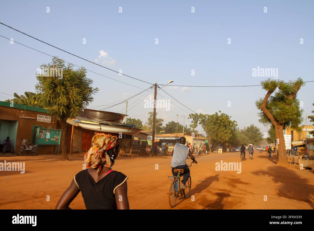 Donna che porta un grande bacino di metallo sulla testa camminando lungo una strada sterrata con un ciclista, Regione del Centro, Ouagadougou, Burkina Faso Foto Stock