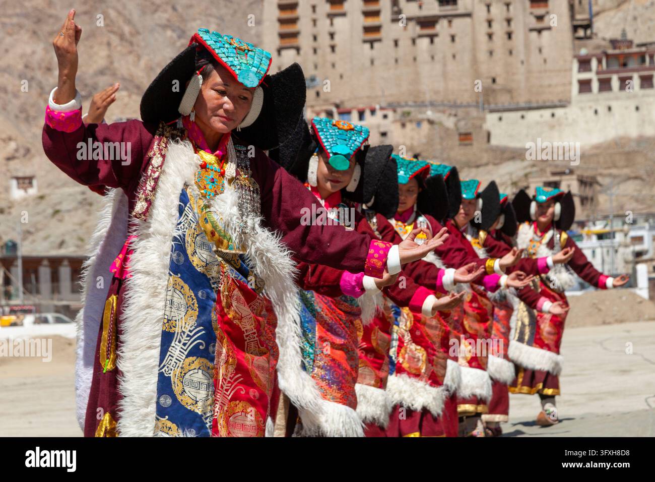 Filiera di donne con copricapi turchesi e mantelli di broccato che eseguono una danza davanti a un palazzo, Ladakh, Leh, India Foto Stock
