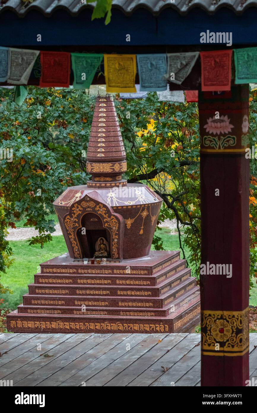 Uno stupa buddista incorniciato da pali di padiglione e bandiere di preghiera si trova in un ambiente di giardino tranquillo che combina artigianato colorato e simbolismo sacro per ispirare Foto Stock