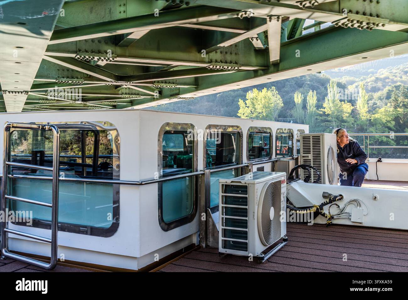 Un ponte d'acciaio verde si estende sul ponte superiore di una nave fluviale sul Douro in Portogallo. Una donna dell'equipaggio è accovacciata sul bordo del ponte accanto a un'unità di climatizzazione, guardando da un lato. Dietro di lei, alberi decidui si ergono sulla collina. Foto Stock