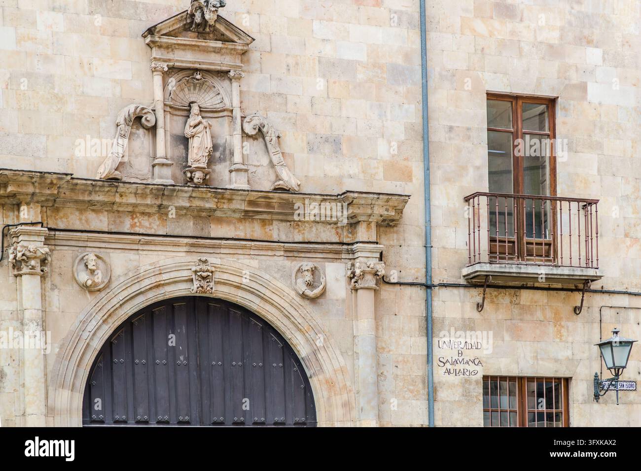 Il portale ad arco tondeggiante di un edificio universitario a Salamanca nella Castiglia e León, in Spagna, è dotato di un portone di legno scuro con borchie. Sopra il cornicione, una figura in pietra si trova in una nicchia a conchiglia sotto un frontone triangolare, tra due volute. Due piccoli medaglioni con teste fiancheggiano l’arco. A destra, sul muro è scritto “UNIVERSIDAD DE SALAMANCA AULARIO” (in inglese: Università di Salamanca, edificio dell’aula magna). Un cartello stradale con la scritta “PLAZA SAN ISIDRO” (in inglese: Piazza San Isidro) pende da una lanterna. Sopra si trova un balcone con una ringhiera in ferro. Foto Stock