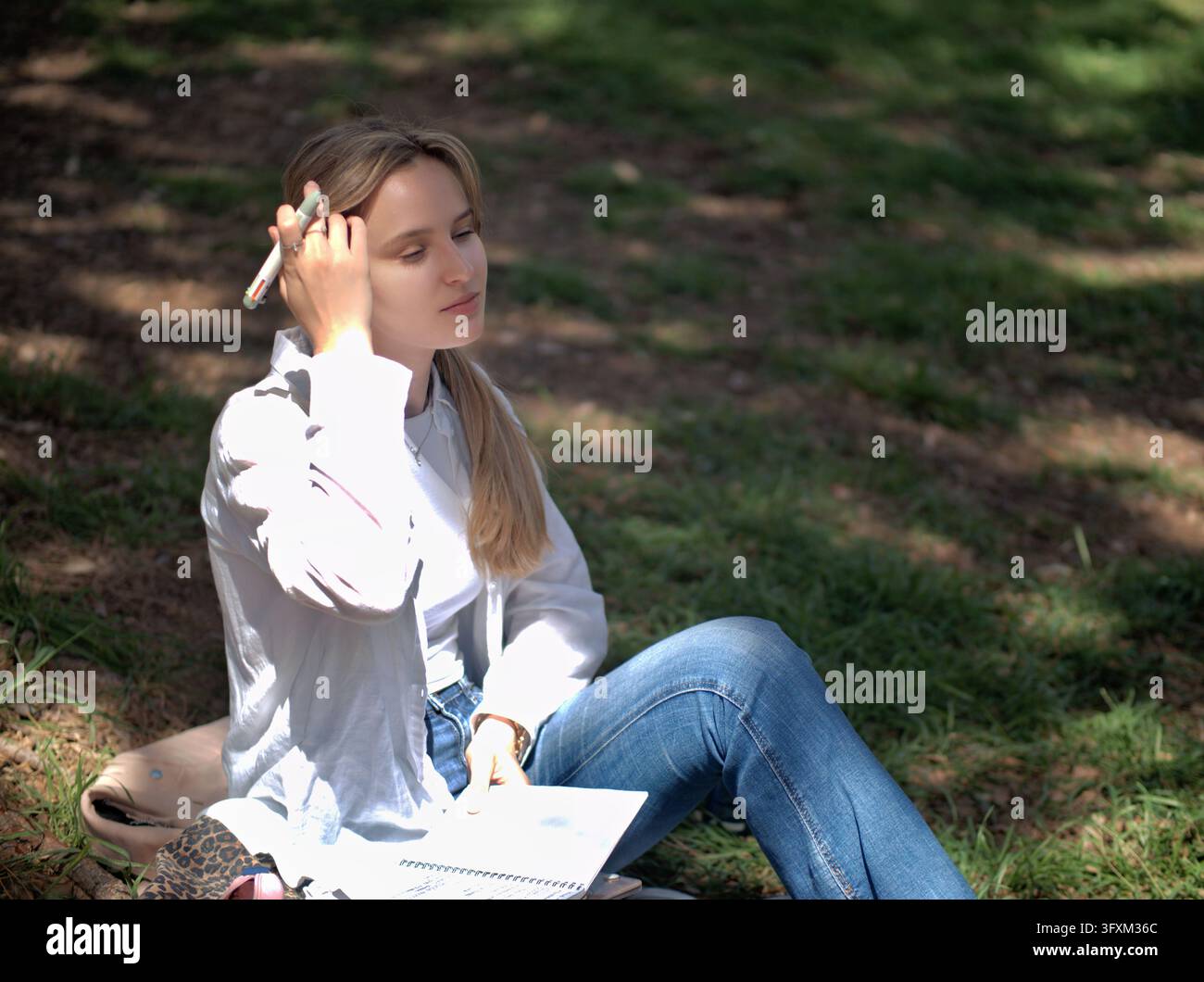 Giovane donna che riposa con gli occhi chiusi seduta sull'erba con un taccuino e una penna in un ambiente di parco all'aperto Foto Stock