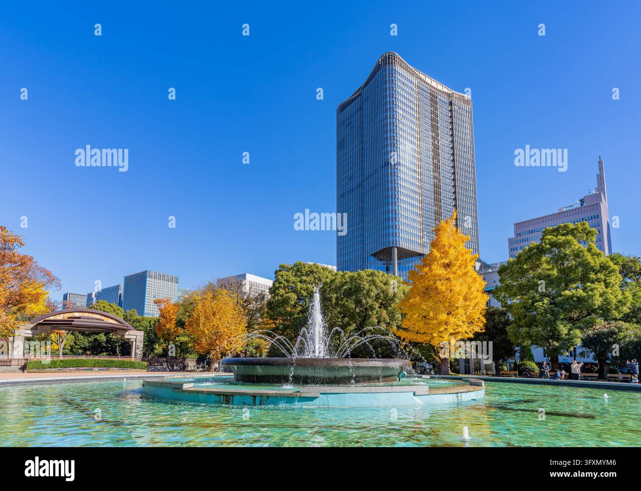 Una fontana d'acqua spruzza in uno stagno del parco circondato da alberi con foglie gialle, sullo sfondo di grattacieli moderni sotto un cielo blu terso. Foto Stock