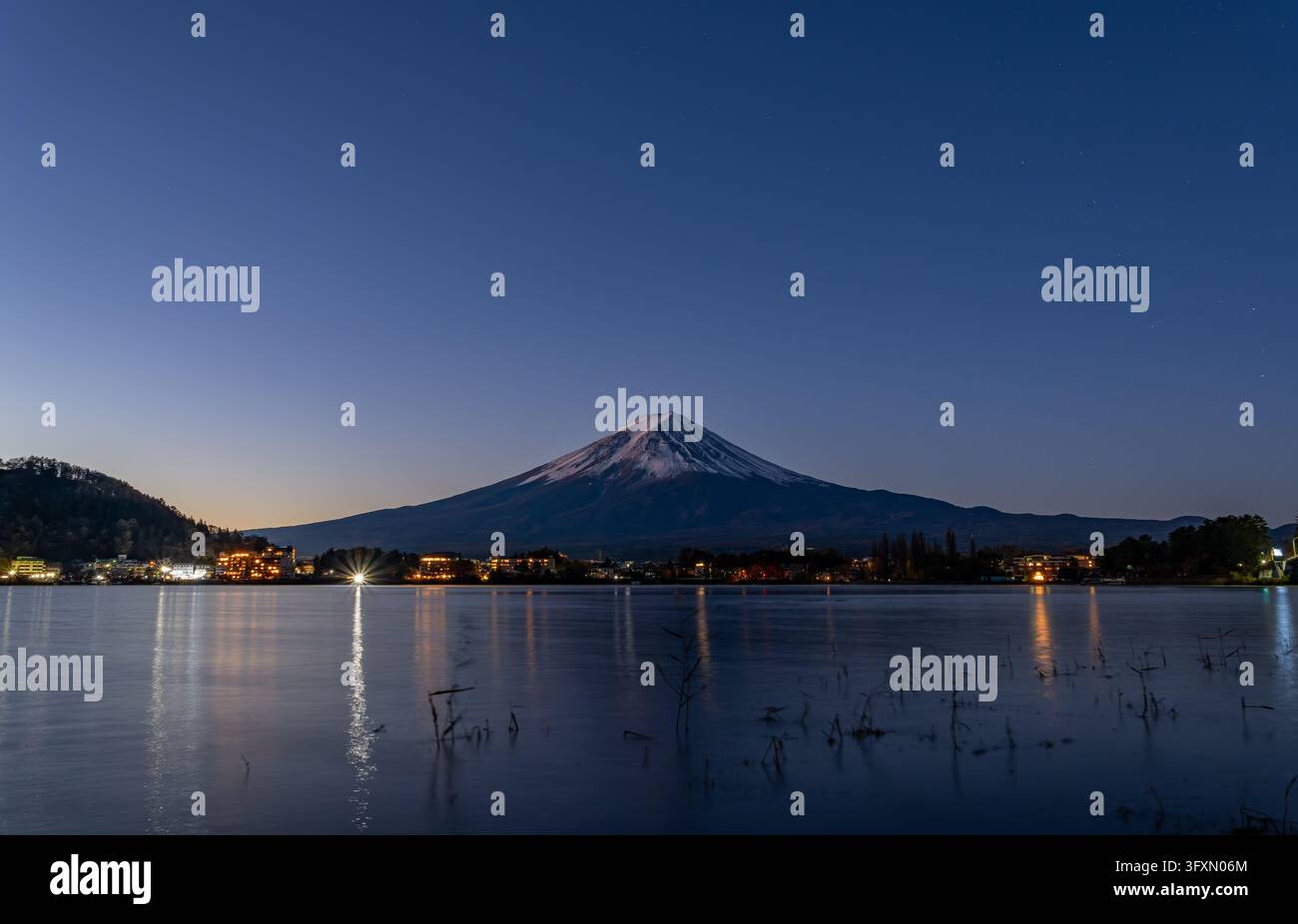 Il monte Fuji coperto di neve si erge sopra una città con edifici illuminati, riflessi nelle acque calme di un lago sotto un cielo notturno stellato. Foto Stock
