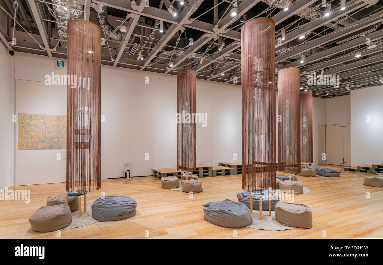Tokyo, Giappone, 22 NOV 2024 - Una spaziosa galleria museale presenta alte installazioni cilindriche con fili e testo giapponese, circondate da pouf Foto Stock