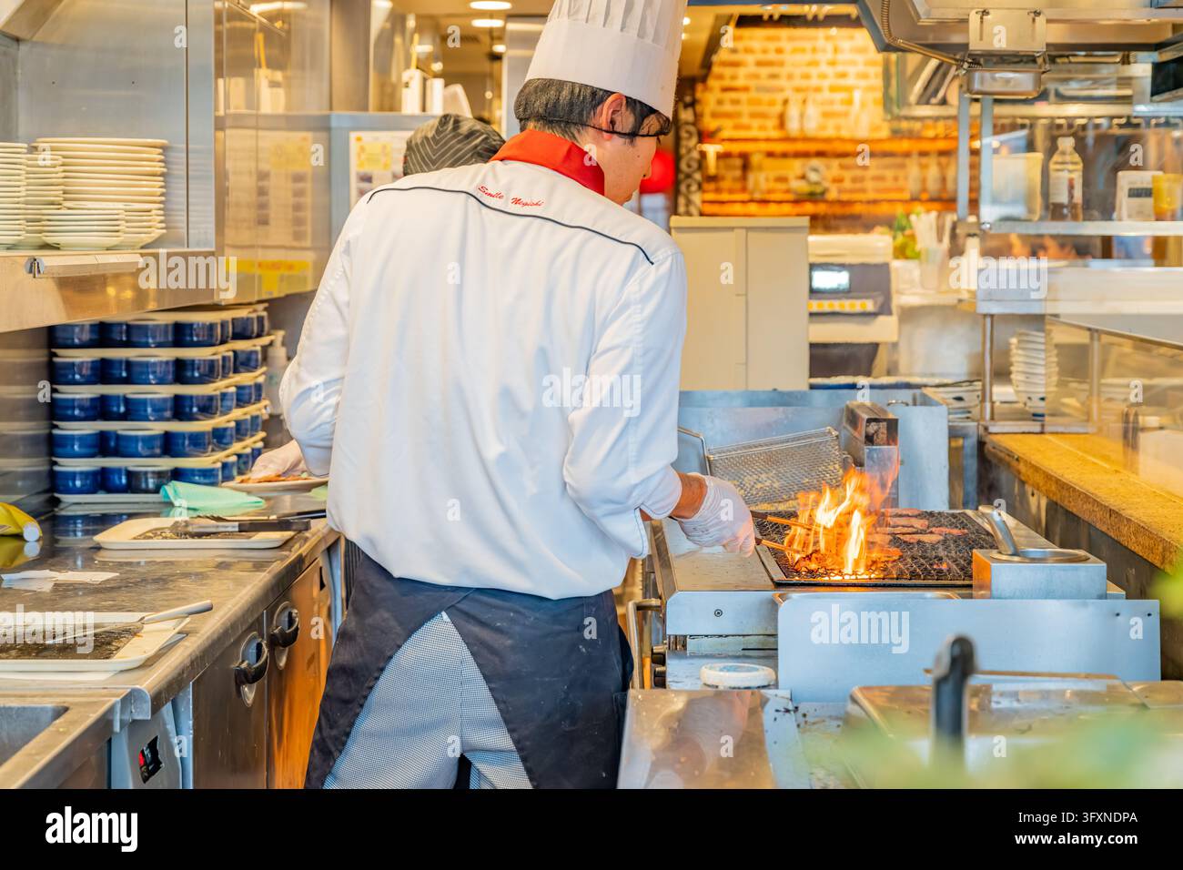 Tokyo, Giappone, 22 NOV 2024 – Uno chef con uniforme e cappello bianchi usa delle pinze per cuocere carne su una griglia fiammeggiante in una cucina di ristorante affollata. Foto Stock