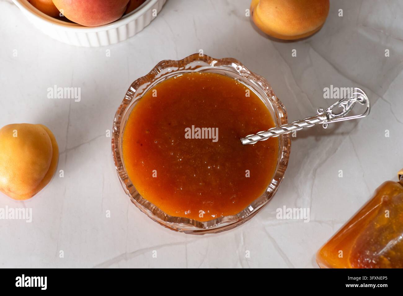 Una vista ravvicinata di marmellata di albicocche fatta in casa in una ciotola di vetro decorativa. Un cucchiaio d'argento riposa nella marmellata, circondato da albicocche fresche e un barattolo di marmellata Foto Stock